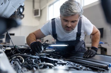 Otomobil tamirhanesinde kaputun altında el feneriyle motoru denetleyen tamirci.