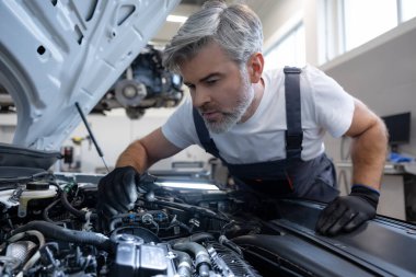 Otomobil tamirhanesinde kaputun altında el feneriyle motor parçalarını inceleyen tamirci.