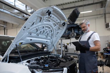 Tamirci otomobil tamirhanesinde motor kaputu açık tabletle teşhis koyuyor.