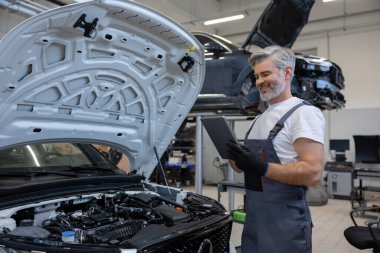 Otomobil tamir atölyesinde elinde tabletle tamirci açık kaputun yanında teşhis koyuyor.