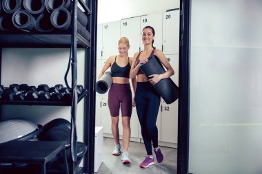 Spor salonunda konuşmak. Spor kıyafetli, sarışın ve esmer kadınlar spor salonunda antremandan önce konuşuyorlar.