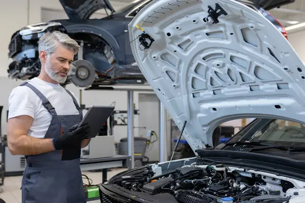 Oto tamir atölyesinde kaputu açık motor kontrolü için tablet kullanan tamirci
