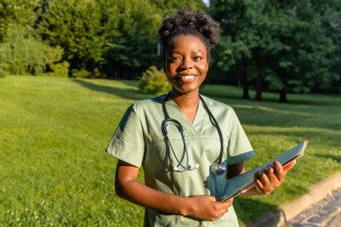 Parkta. Yeşil arka planda laboratuvar önlüklü, koyu tenli gülümseyen bir kadın.