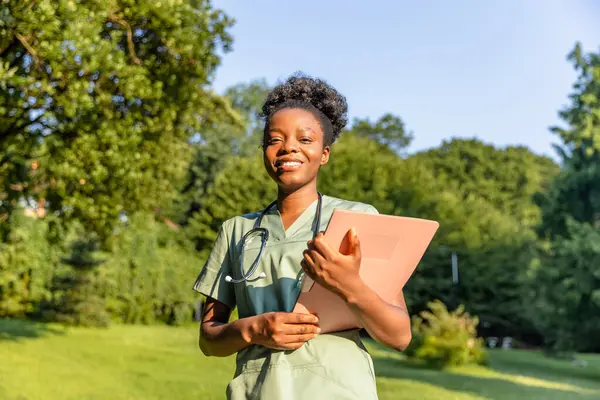 Parkta. Yeşil arka planda laboratuvar önlüklü, koyu tenli gülümseyen bir kadın.
