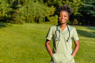 Parktaki hemşire. Parkta boynunda laboratuvar önlüğü ve steteskop olan Afrikalı Amerikalı genç bir kadın.