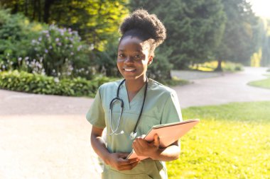 Parkta. Yeşil arka planda laboratuvar önlüklü, koyu tenli gülümseyen bir kadın.