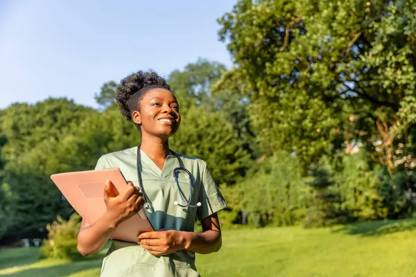 Parkta. Yeşil arka planda laboratuvar önlüklü, koyu tenli gülümseyen bir kadın.