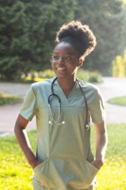 Parktaki hemşire. Parkta boynunda laboratuvar önlüğü ve steteskop olan Afrikalı Amerikalı genç bir kadın.