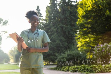 Parkta. Yeşil arka planda laboratuvar önlüklü, koyu tenli gülümseyen bir kadın.