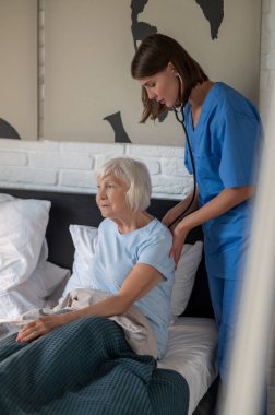 Bakım evi. Doktor yaşlı hastayı muayene ediyor ve konsantre görünüyor.