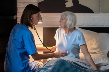 Bakım evi. Doktor yaşlı hastayı muayene ediyor ve konsantre görünüyor.