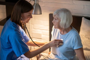 Bakım evi. Doktor yaşlı hastayı muayene ediyor ve konsantre görünüyor.