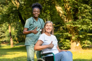 Güzel yürüyüş. Tekerlekli sandalyedeki kadın, parkta bir hemşireyle yürürken mutlu görünüyor.