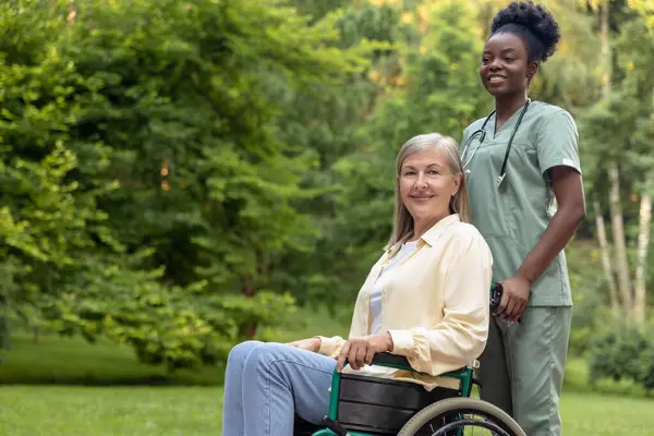 İyileşme. Afrikalı Amerikalı hemşire, parkta tekerlekli sandalyeyle kadın hasta gezdiriyor.