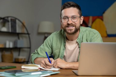 Evde dizüstü bilgisayarda yazan sakallı bir adam. Masada oturmuş, defter açık, kalem de rahat bir odada.