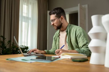 Sakallı adam, raporları analiz ediyor. Masada dizüstü bilgisayarla oturuyor ve rahat bir ortamda belgeler hazırlıyor.