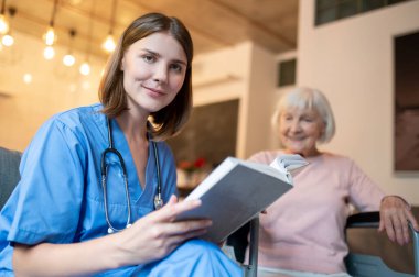 Birlikte okuyoruz. Yaşlı bir bayana kitap okuyan bir kadın bakıcı.