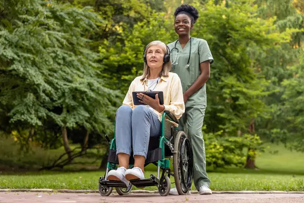 Sabah yürüyüşü. Tekerlekli sandalyedeki kadın ve hemşire parkta yürüyor.