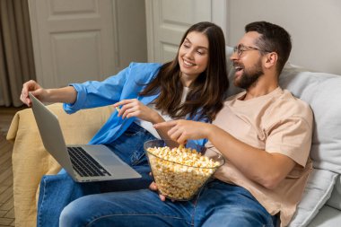 Gülümseyen adam ve kadın dizüstü bilgisayarda film izlerken kanepede sarılıp evde boş vakitlerin tadını çıkarıyorlar.