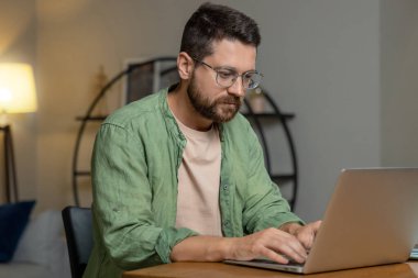 Sakallı adam sunum hazırlıyor. Masada dizüstü bilgisayarla oturuyor ve rahat bir odada kayıyor.