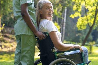 Güzel yürüyüş. Tekerlekli sandalyedeki kadın, parkta bir hemşireyle yürürken mutlu görünüyor.