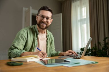 Sakallı adam, raporları analiz ediyor. Masada dizüstü bilgisayarla oturuyor ve rahat bir ortamda belgeler hazırlıyor.