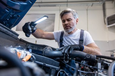 Otomobil tamirhanesinde kaputun altında el feneriyle motoru denetleyen tamirci.