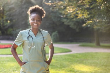 Parktaki hemşire. Parkta boynunda laboratuvar önlüğü ve steteskop olan Afrikalı Amerikalı genç bir kadın.