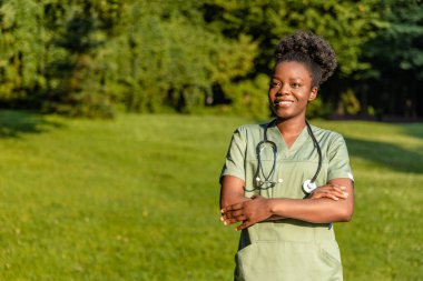 Parktaki hemşire. Parkta boynunda laboratuvar önlüğü ve steteskop olan Afrikalı Amerikalı genç bir kadın.