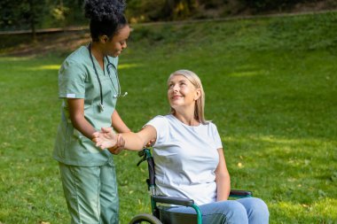 Güzel yürüyüş. Tekerlekli sandalyedeki kadın, parkta bir hemşireyle yürürken mutlu görünüyor.