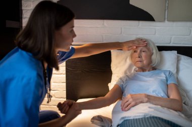 Ateşim var. Doktor alnına dokunarak yaşlı bir kadının ateşini ölçüyor.