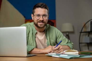 Evde dizüstü bilgisayarda yazan sakallı bir adam. Masada oturmuş, defter açık, kalem de rahat bir odada.