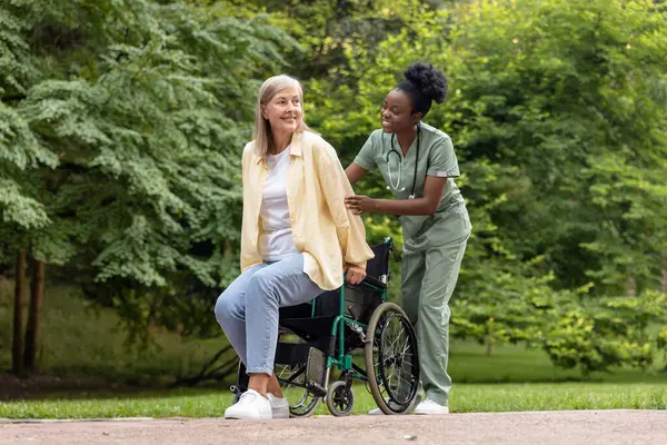 İyileşme. Afrikalı Amerikalı hemşire, parkta tekerlekli sandalyeyle kadın hasta gezdiriyor.