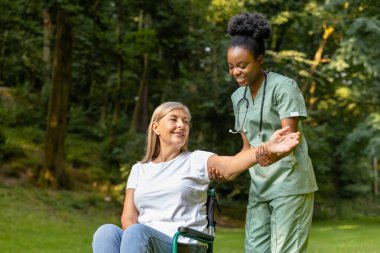 Güzel yürüyüş. Tekerlekli sandalyedeki kadın, parkta bir hemşireyle yürürken mutlu görünüyor.