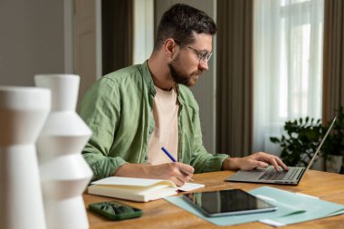 İç işlerinde bilgisayar ve kağıtlarla çalışırken masadaki notları inceleyen sakallı adam.