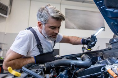 Otomobil tamirhanesinde kaputun altında el feneriyle motoru denetleyen tamirci.