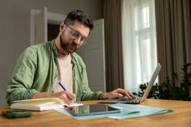 Sakallı adam ofisteki bilgisayarda not alıyor. Masada oturuyor. Elinde defter ve kalemle sessiz bir ortamda.