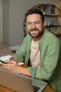 Ofiste çalışan sakallı bir adam rahat bir yerde dizüstü bilgisayarı ve akıllı telefonu olan bir masada oturuyor.