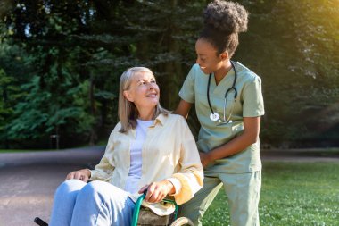 Parkta yürü. Afrikalı Amerikalı hemşire, parkta kadın bir hastayla tekerlekli sandalyeyi yuvarlıyor.