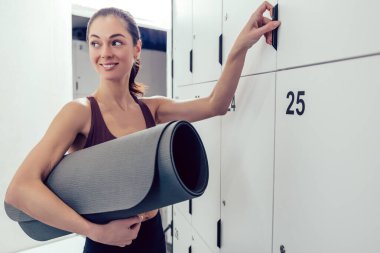 Spor salonunda. Siyah saçlı genç bir kadın spor salonundaki soyunma odasında dolabı açıyor.