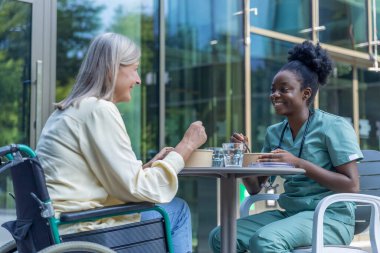 Öğle yemeği vakti. Hemşire ve bayan hasta kliniğin bahçesinde yemek yiyorlar ve ilgili görünüyorlar.