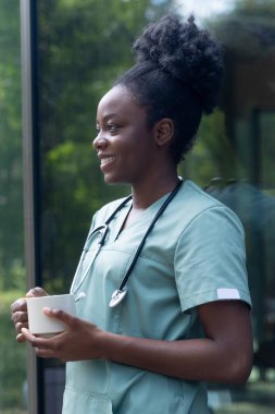 Kahve zamanı. Laboratuvar önlüklü Afrikalı Amerikalı kadın elinde bir fincan kahveyle kliniğin kapısında duruyor.