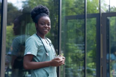 Kahve zamanı. Laboratuvar önlüklü Afrikalı Amerikalı kadın elinde bir fincan kahveyle kliniğin kapısında duruyor.