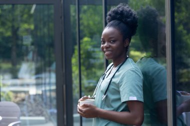 Kahve zamanı. Laboratuvar önlüklü Afrikalı Amerikalı kadın elinde bir fincan kahveyle kliniğin kapısında duruyor.