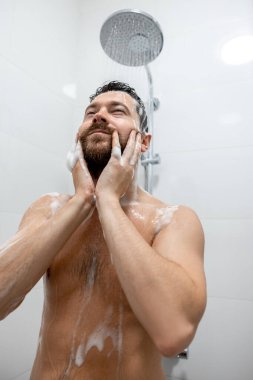 Esmer adam, beyaz banyoda şampuanla başını yıkıyor.