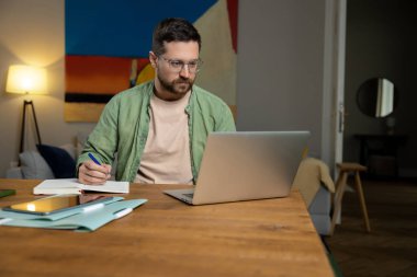 Odaklanmış, gözlüklü, yeşil tişörtlü, dizüstü bilgisayarlı, tahta masa örtüsü oturma odasında oturan bej tişörtlü adam.