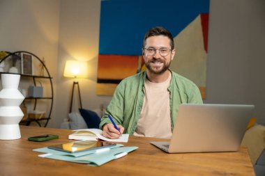 Gözlüklü, yeşil tişörtlü, dizüstü bilgisayarlı, not defterli, ofis ortamındaki ahşap masada oturan genç bir serbest çalışan.