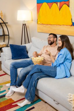 Gözlüklü, bej gömlekli, uzun saçlı, mavi gömlekli, patlamış mısır yiyen, rahat bir evde televizyon izleyen bir adam.