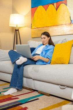 Rahat kadın serbest giyinen, dizüstü bilgisayar kullanan, şık oturma odasındaki, soyut duvar resimli yumuşak koltukta oturan biri.