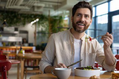 Restoran masasında salata yiyen sakallı adam modern yemek ortamında çatal ve kâseyle.